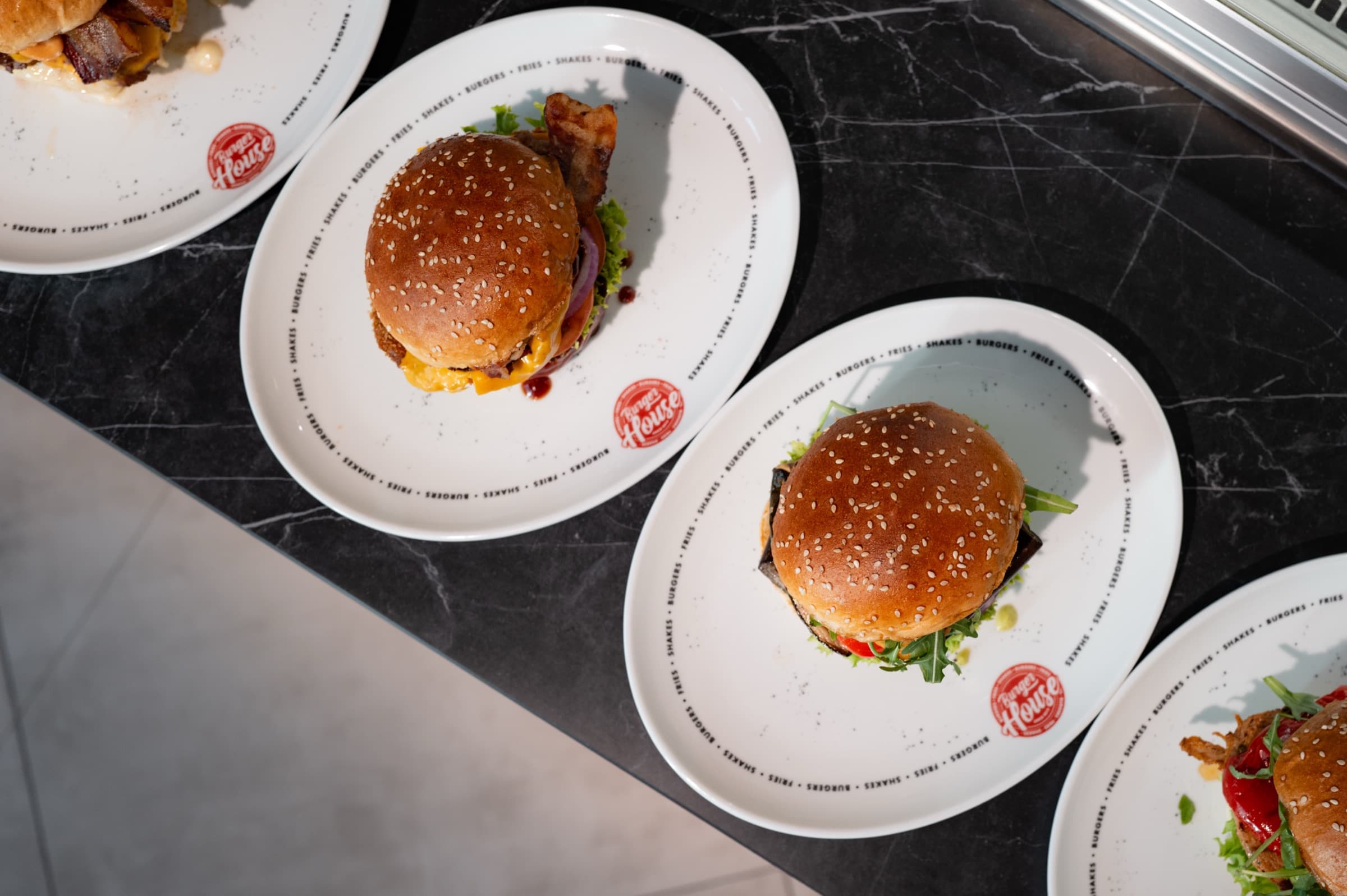 Burger House Burger auf weißen Tellern mit Logo, von oben fotografiert: Sesambrötchen, Bacon, Cheddar und Rucola auf dunklem Marmor